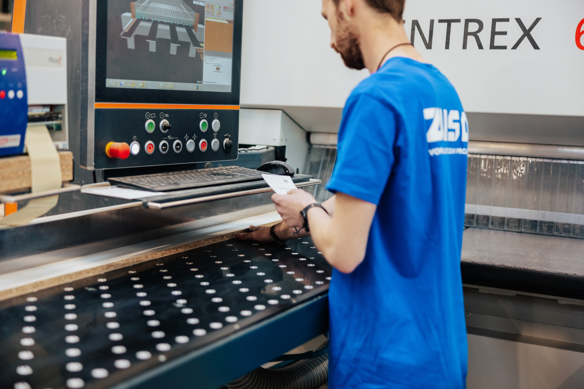 Ein Mann in blauem T-Shirt steht vor einer CNC-Maschine, liest ein Blatt Papier und arbeitet an einem Bedienfeld.