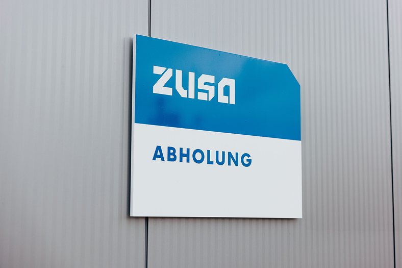 Ein Schild mit der Aufschrift „ZUSA Abholung“ in blau-weißer Farbe, montiert an einer geriffelten Metallwand.
